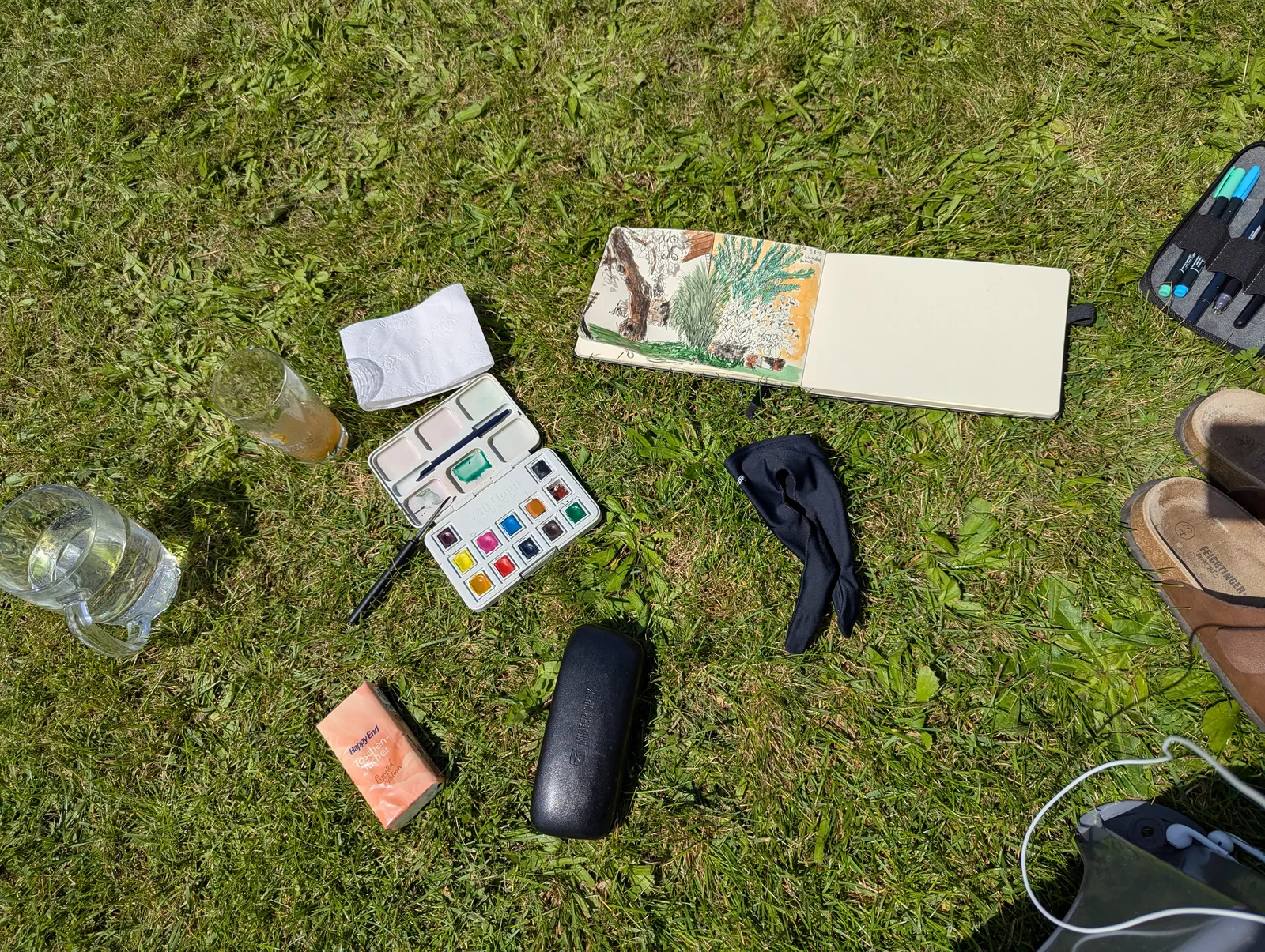 Watercolor supplies laid out in the grass, sketchbook open to an in-progress garden drawing